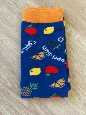 Capri-Sun strumpor - Säljer ett par blåa strumpor med färgglada fruktmotiv som citron, apelsin, ananas, vindruvor och äpple. Strumporna har orange mudd och texten 'Capri Sun' insprängd i mönstret. Perfekta för dig som gillar lekfulla detaljer i din outfit.