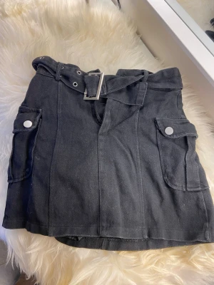 Svart kortkjol med bälte från FItS - Snygg svart kortkjol från FItS med bälte i midjan och stora fickor med knappar på sidorna. Kjolen har en rak passform och är gjord i ett kraftigare bomullstyg. Perfekt för dig som gillar utility-stil och vill ha något trendigt i garderoben.