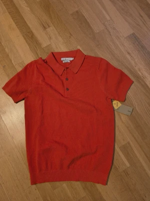 Orange Pike från S.C.W - Kortärmad pike från S.C.W med klassisk krage och tre knappar framtill. Toppen är tillverkad i ett stickat material och har en ribbad nederkant. Perfekt för dig som vill ha en stilren och bekväm look.