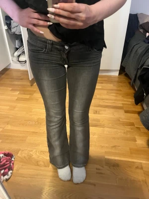 Grå bootcut jeans med låg midja - Säljer ett par grå bootcut jeans med låg midja och klassisk femficksmodell. Jeansen har en snygg tvättad look och är tillverkade i stretchigt denimtyg för skön passform. Perfekta för dig som gillar en retroinspirerad stil. storlek 27/30