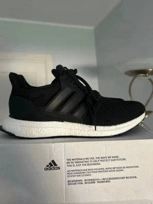 Adidas UltraBoost svart sneaker - Svarta Adidas UltraBoost sneakers med stickad mesh-överdel och klassiska tre ränder på sidan. Skon har en vit Boost-mellansula och svart yttersula. Låg modell med snörning och bekväm passform, perfekt för dig som gillar stil och komfort.