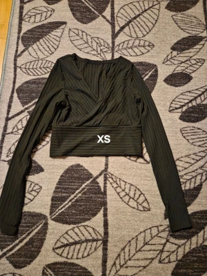 Svart omlottopp med ribbad struktur - Trendig svart omlottopp med ribbad struktur och croppad passform. Toppen har långa ärmar och bred linning nedtill. Perfekt för dig som gillar stilrena och moderna plagg.