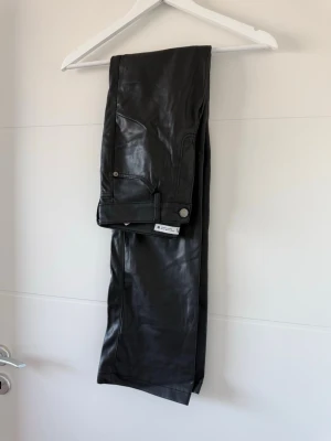 Svarta coated jeans från Zara - Säljer ett par svarta coated jeans från Zara med fem fickor och raka ben. Jeansen har en snygg glansig finish som ger en cool läderlook och stängs med knapp och dragkedja. Perfekta för dig som vill ha en edgy stil.