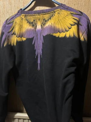 Marcelo Burlon - Unik svart sweatshirt från Marcelo Burlon med ett stort tryck av gula och lila vingar över axlar och bröst. Tröjan har rund halsringning och långa ärmar. Perfekt för dig som vill sticka ut med en cool och konstnärlig design.