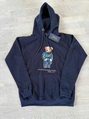 Marinblå Polo Ralph Lauren hoodie - Snygg marinblå hoodie från Polo Ralph Lauren med den ikoniska Polobear-figuren tryckt på bröstet. Hoodien har huva med dragsko, magficka och ribbade muddar. Tillverkad i mjukt bomullsmaterial som är skönt att bära.