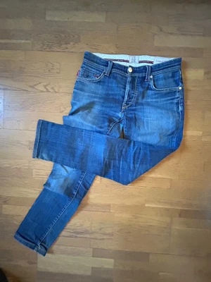 Blå jeans från tramarosa  - Säljer ett par klassiska blå jeans från Tramarosa med raka ben och fem fickor. Jeansen har snygga slitningar och kontrastsömmar samt detaljer i rött vid fickorna. Tillverkade i bomull med en skön stretch för extra komfort. Det billiga priset beror på ett litet hål på byxornas underdel