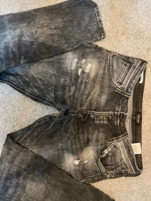 Svarta slitna jeans från Jack & Jones - Säljer ett par svarta jeans från Jack & Jones med snyggt slitna detaljer och tvättad look. Jeansen har klassisk femficksmodell, dragkedjegylf och raka ben. Materialet är mjukt denim som ger en avslappnad vibe.