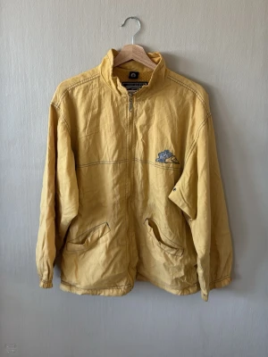 Gul vindjacka från Quiksilver - Snygg gul vindjacka från Quiksilver med dragkedja framtill och två snedställda fickor. Jackan har meshfoder, hög krage och en broderad logga på bröstet. Perfekt för dig som gillar streetwear och vill sticka ut med en retrostil.