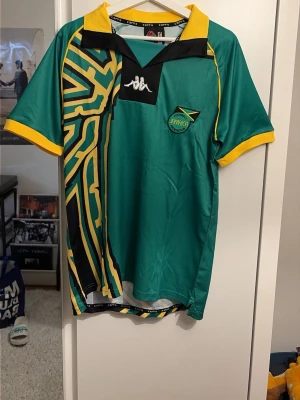 Jamaica matchtröja från Kappa - Snygg grön matchtröja från Kappa med Jamaicas landslagsmärke på bröstet. Tröjan har gula och svarta detaljer, grafiskt mönster på ena sidan och gul krage samt ärmslut. Tillverkad i lätt och ventilerande polyester, perfekt för sport och träning.