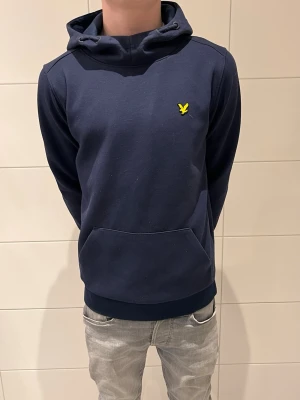 Mörkblå hoodie från Lyle & Scott - Mörkblå hoodie från Lyle & Scott med klassisk gul logga på bröstet. Hoodien har känguruficka framtill och ribbade muddar vid ärmar och nederkant. Tillverkad i mjuk bomull och har en avslappnad passform.