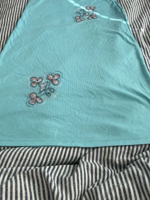 Ljusblå ribbad klänning med broderi - Säljer en ljusblå ribbad klänning med broderade blommor i rosa och vitt framtill. Klänningen har en enkel, rak passform och är ärmlös. Perfekt för varma dagar och ger en söt och fräsch look.