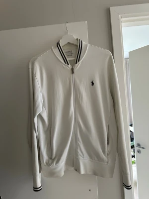 Vit zip hoodie från Polo Ralph Lauren - Snygg vit hoodie med dragkedja från Polo Ralph Lauren. Klassisk design med svarta ränder på krage och muddar. Bra skick. Storlek S.