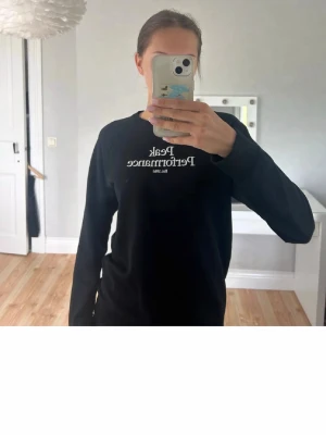 Svart långärmad topp från Peak Performance - Svart långärmad topp från Peak Performance med vit logga och text på bröstet. Klassisk rund halsringning och enkel, stilren design som passar till det mesta. Perfekt för dig som gillar en clean och sportig look.