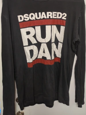 Svart långärmad t-shirt Dsquared2 - Cool svart långärmad t-shirt från Dsquared2 med stort vitt och rött tryck på framsidan där det står 'RUN DAN'. Skön passform och tillverkad i mjuk bomull. Perfekt för dig som gillar streetwear och vill sticka ut med ett statement-plagg.