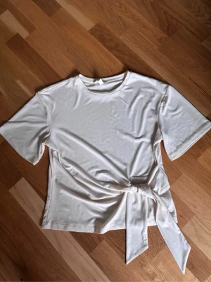 Vit topp med knyt i midjan - Stilren vit topp med rund halsringning och korta ärmar. Toppen har en snygg knyt-detalj i midjan som ger en trendig touch. Perfekt att matcha med jeans eller kjol för en enkel men chic look.