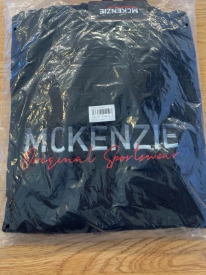 McKenzie hoodie - Svart hoodie från McKenzie med stort tryck på bröstet där det står 'McKenzie Original Sportswear' i vitt och rött. Klassisk modell med huva och sportig känsla, perfekt för dig som gillar streetwear och enkel stil. Hör av er om ni vill se mer bilder råkade köpa 2 för ett tag sen och endast öppnat en.😊