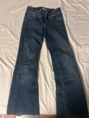 Blå bootcut jeans med dubbla knappar - Säljer ett par blå bootcut jeans med dubbla knappar i midjan och vita kontrastsömmar. Jeansen har klassiska fem fickor och coola broderade detaljer på bakfickorna. Perfekta för dig som gillar retrovibbar och en avslappnad stil.
