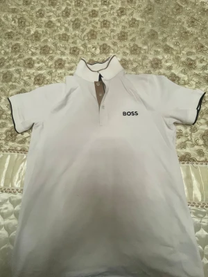 Vit kortärmad t-shirt från BOSS - Vit t-shirt från BOSS med korta ärmar och klassisk krage. T-shirten har svarta detaljer på kragen och ärmsluten samt BOSS-logga broderad på bröstet. Knappslå framtill med kontrastfärgade knappar. Perfekt för en clean och stilren look.