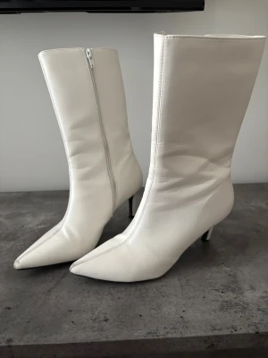 Vita spetsiga boots med klack - Säljer ett par vita boots med spetsig tå och smal klack. Stövlarna har dragkedja på insidan och är tillverkade i ett slätt material som ser ut som skinn. De har en stilren och trendig look som passar till många outfits.