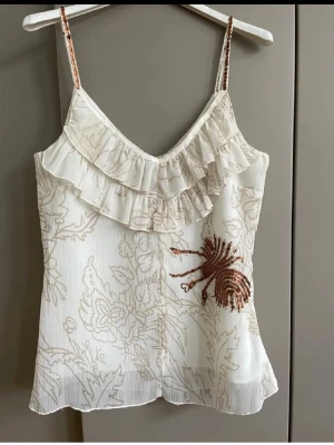  linnetopp med volang och paljetter ny med prislappen  - Söt  linnetopp med tunna axelband, volangdetalj framtill och diskret blommigt mönster. Toppen har en dekorativ paljettapplikation i kopparfärg på ena sidan och är perfekt för sommardagar. Materialet känns lätt och luftigt.