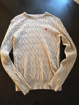 Beige kabelstickad tröja Morris - Snygg beige kabelstickad tröja från Morris med rund halsringning och diskret röd logga broderad på bröstet. Tröjan har långa ärmar och ribbade muddar vid ärmslut och nederkant. Perfekt för dig som gillar klassisk och stilren design. Köpt på Åhléns nypris 1500kr