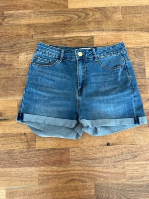 Blå denimshorts med uppvik - Snygga blå denimshorts med uppvikta benslut och klassisk femficksdesign. Shortsen har normal passform, knapp och dragkedja framtill samt bälteshällor. Perfekta för varma dagar och enkla att matcha med olika stilar.