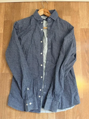 Blå mönstrad skjorta J.Lindeberg - Säljer en blå slim fit skjorta från J.Lindeberg med små vita prickar. Skjortan är 100% bomull och sitter riktigt skönt. Inga defekter. Tveka inte på att höra av dig ifall du undrar något!✨