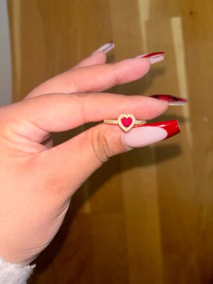Guldfärgad ring med hjärtformad sten - Snygg guldfärgad ring med en hjärtformad röd sten i mitten, omgiven av små gnistrande stenar runt hjärtat och längs ringen. Perfekt för dig som vill ha en romantisk och trendig accessoar som sticker ut.