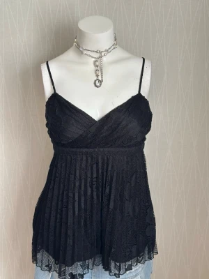 Svart spetsig linnetopp med tunna band - Snygg svart linnetopp med tunna axelband och omlottskärning framtill. Toppen är gjord i spets med plisserad nederdel och har en luftig, flowy passform. Perfekt för att lyfta vilken outfit som helst med en elegant touch.