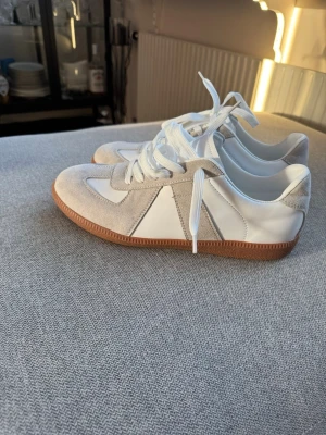 Vita och grå sneakers med mockadetaljer GATS - Vita Gats sneakers i vitt och ljusgrått med ovandel i läder och mocka. Skorna har klassisk gummisula i brunt och vita skosnören. Perfekta för dig som gillar minimalistisk och clean stil. Passar till jeans eller shorts för en avslappnad look. Storlek 44 brand new