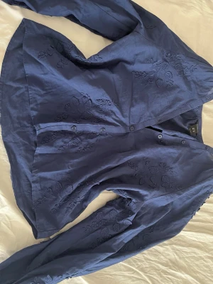 Mörkblå broderad blus  - Snygg mörkblå blus med diskreta blombroderier över hela tyget. Blusen har knäppning framtill och långa ärmar. Perfekt för dig som gillar en stilren men ändå detaljrik look. Materialet känns lätt och luftigt.