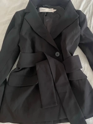 Svart kavaj med skärp från MQ - Stilren svart kavaj från MQ med breda slag, ett knäppningsknapp framtill och matchande tygskärp i midjan. Kavajen har två fickor med lock och är tillverkad i ett slätt tyg som ger en modern look. Perfekt för dig som gillar minimalistisk stil.