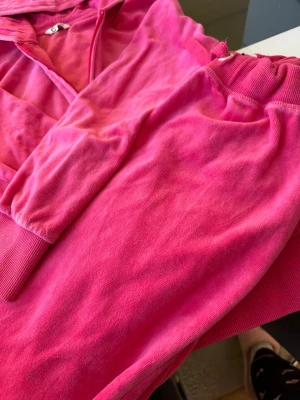 Rosa velourset med hoodie och byxor - Mysigt loungewear-set i starkt rosa velour. Setet består av en hoodie med dragkedja och huva samt matchande byxor med resår i midjan. Perfekt för chill dagar hemma eller när du vill vara extra bekväm.