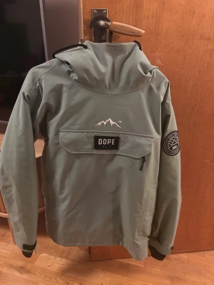 Grön DOPE anorak med huva - Säljer en grön anorak från DOPE med stor ficka framtill, huva med dragsko och svart logga på bröstet. Jackan har en patch på ärmen och är tillverkad i slitstark polyester. Perfekt för utomhusaktiviteter och streetwear.