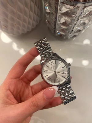 Silverfärgad klocka från Zone - Stilren klocka från Zone med rund urtavla i silver och romerska siffror. Armbandet är i metall med klassisk länkdesign och hela klockan har en elegant, blank finish. Perfekt accessoar för dig som gillar en tidlös look.