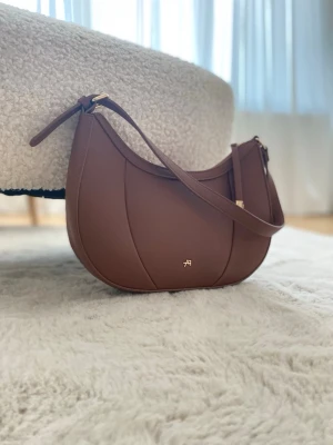 Brun axelväska från AH - Snygg brun axelväska från AH i halvmåneformad design. Väskan är gjord i slätt läder och har ett justerbart axelband samt guldfärgade metalldetaljer. Liten logga framtill. Perfekt för dig som gillar stilrena accessoarer.