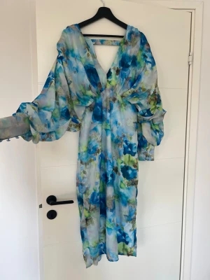 Blommig långklänning med v-ringning - Säljer en flowy långklänning med v-ringning och vida ärmar. Klänningen har ett blommigt mönster i blått, grönt och vitt och är tillverkad i ett tunt, lätt material som ger en luftig känsla. Perfekt för sommarens alla tillfällen!
