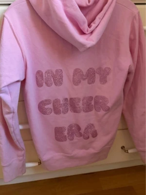 Rosa hoodie med glittertext - Snygg ljusrosa hoodie med huva och magficka. På ryggen står det 'IN MY CHEER ERA' i stora, glittriga bokstäver. Hoodien har dragsko i huvan och ribbade muddar vid ärmar och nederkant. Perfekt för dig som gillar trendiga och bekväma plagg. Skulle säga att den är liten k storlek så passar även xs beroende på hur man vill ha den 