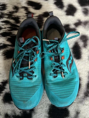 Turkosa Saucony sneakers mesh - Säljer ett par turkosa sneakers från Saucony med svart logga på sidan. Skorna har ovandel i ventilerande mesh, mönstrade skosnören och detaljer i orange. Låg modell med vadderad häl och tå för extra komfort. Perfekta för dig som gillar sportig stil.