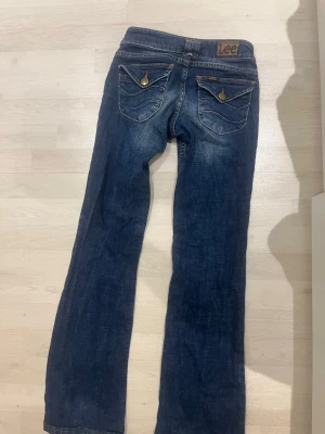 Mörkblå bootcut jeans från Lee - Säljer ett par mörkblå bootcut jeans från Lee med snygga slitningar och kontrastsömmar. Jeansen har två bakfickor med lock och knappdetaljer samt klassisk Lee-lapp i midjan. Tillverkade i denim med en skön passform.