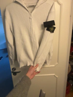 Vit stickad ziptröja Stone Island - Vit stickad ziptröja från Stone Island med ribbad struktur och huva. Tröjan har hel dragkedja framtill och den klassiska Stone Island-loggan på ärmen. Perfekt för lager-på-lager och streetwear-vibe.