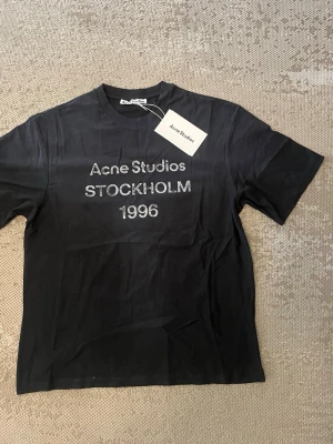 Svart t-shirt Acne Studios Stockholm  - Svart t-shirt från Acne Studios med tryck 'Acne Studios STOCKHOLM 1996' på bröstet. Klassisk rund hals och korta ärmar. Materialet är mjuk bomull och passformen är normal. Perfekt för dig som gillar stilrena och trendiga plagg.