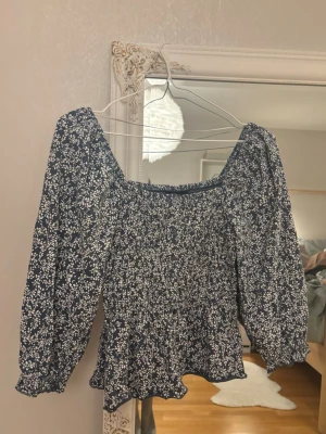 Blommig blus med puffärm - Supersöt blommig blus i mörkblått och vitt med smockad midja och volangkant. Blusen har långa puffärmar och en fyrkantig urringning samt knapp detalj. Perfekt för dig som gillar romantisk stil och vill ha något som sticker ut lite extra.