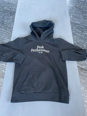 Svart hoodie från Peak Performance - Snygg svart hoodie från Peak Performance med vit broderad logga och text 'Est. 1986' på bröstet. Klassisk känguruficka framtill och ribbade muddar vid ärmslut och nederkant. Perfekt för en avslappnad och sportig stil. Kom med minsta lilla fundering! 😁