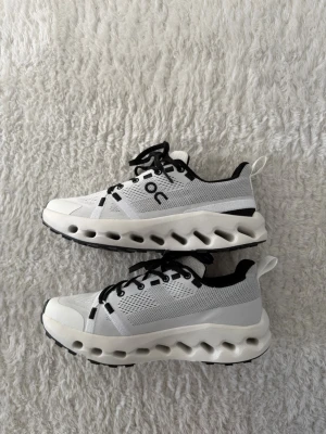 Helt nya vita On cloud sneakers med svarta detaljer - Helt nya! Snygga vita sneakers från On cloud med svarta detaljer och snörning. Ovandelen är i mesh för bra ventilation och sulan har On's karakteristiska dämpning. Perfekta för dig som gillar sportig och modern stil.