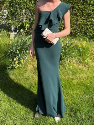 Mörkgrön one shoulder långklänning - Elegant mörkgrön långklänning med volangdetaljer och oneshoulder-design från Lulus. Klänningen har en figurnära passform och är perfekt för speciella tillfällen. Materialet är slätt och följsamt, vilket ger en lyxig känsla. Perfekt till studentbal!