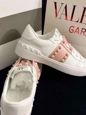 Valentino Garavani vita sneakers - Snygga vita sneakers från Valentino Garavani med ljusrosa band och ikoniska nitar i guldton. Skorna har en låg silhuett, rund tå och är tillverkade i slätt läder. Perfekta för dig som vill ha en exklusiv och trendig look.