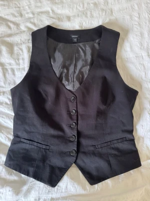 Svart väst med knappar - Klassisk svart väst med fem knappar framtill och två dekorativa fickor. Västen har v-ringad hals och är tillverkad i ett slätt tyg som ger en stilren look. Perfekt att styla över en skjorta för en trendig outfit. Västen är i storlek 42 men passar mig som är 36-38 eftersom man kan dra in i ryggen, därav också har jag angivit 38 som storlek!🤍