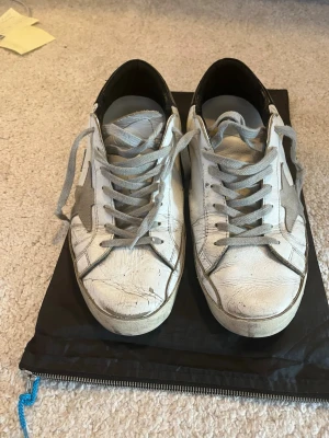 Vita Golden Goose sneakers med grå stjärna - Snygga vita sneakers från Golden Goose med den ikoniska grå stjärnan på sidan och grå skosnören. Skorna har en klassisk låg siluett och är tillverkade i läder med slitna detaljer för en cool, avslappnad look. Yttersulan är i gummi och har fiskbensmönster.