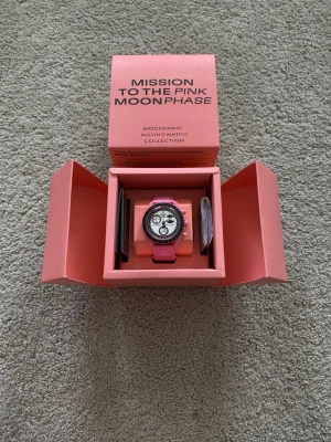 Omega x Swatch Pink Moonphase klocka - Säljer en Omega x Swatch MoonSwatch Pink Moonphase med rosa biokeramisk boett och matchande rosa armband. Klockan har vit urtavla med svarta och rosa detaljer samt kronograf och månfaskomplication. Kommer i originalförpackning.